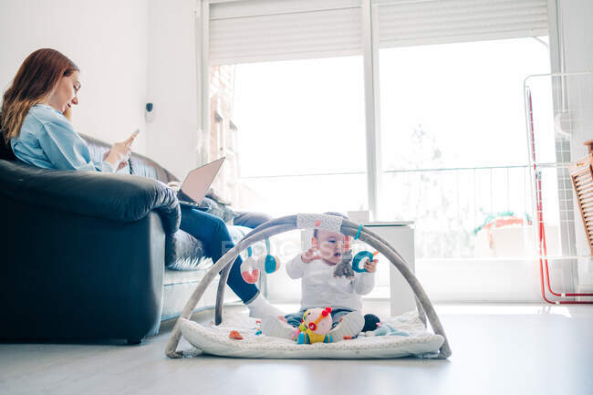 Focada jovem mãe em roupas casuais navegando no smartphone e netbook sentado no sofá perto adorável bebê brincando com brinquedos no chão na sala de estar — Fotografia de Stock