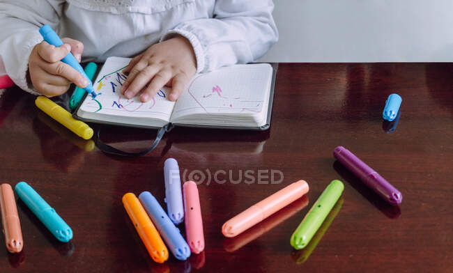 Crop unrecognizable little child sitting at table and drawing with markers in notebook — Stock Photo
