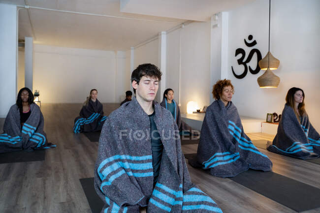 Мультирасовые спокойные люди, завернутые в пледы, сидят на ковриках в студии и медитируют во время занятий йогой — стоковое фото