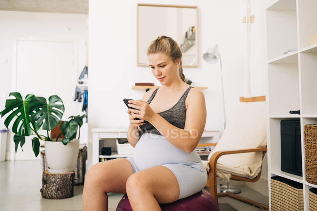 Jovem expectante conteúdo feminino sentado na bola de fitness enquanto navega smartphone em casa — Fotografia de Stock