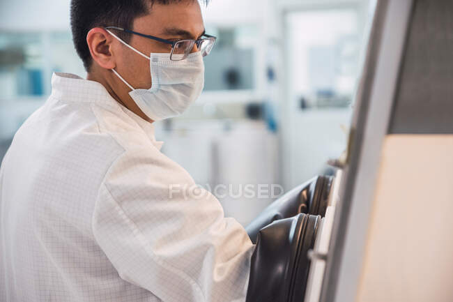 Side view crop concentrated male scientist in lab coat and mask holding hands in biosafety cabinet while working with contaminated materials — Stock Photo