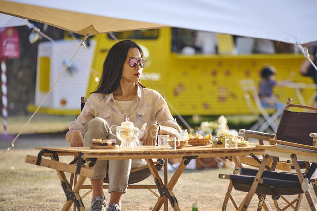 Beautiful ethnic Asian female in sunglasses sitting at table while having a relaxing time in camping area during holidays looking away — Stock Photo