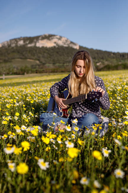 Young hipster woman sitting on a meadow in the countryside playing guitar during summer sunlight. — Stock Photo