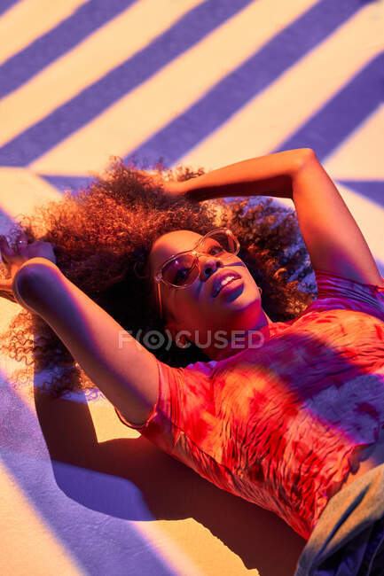 From above attractive young African American female in trendy sunglasses lying gracefully on floor in neon lights and looking at camera — Stock Photo