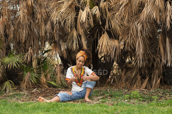 Contenido joven hembra negra en ropa ornamental sentada en el prado contra palmeras con hojas descoloridas - foto de stock