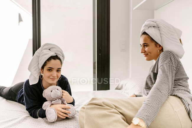 Happy young ethnic female best friends in casual clothes and towels on heads relaxing on comfortable bed after having bath at home — Photo de stock
