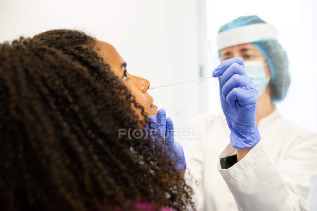 Specialista di medicina femminile in uniforme protettiva, guanti in lattice e maschera facciale facendo test del tampone nasale con batuffolo di cotone sulla paziente afroamericana in clinica durante l'epidemia di coronavirus — Foto stock