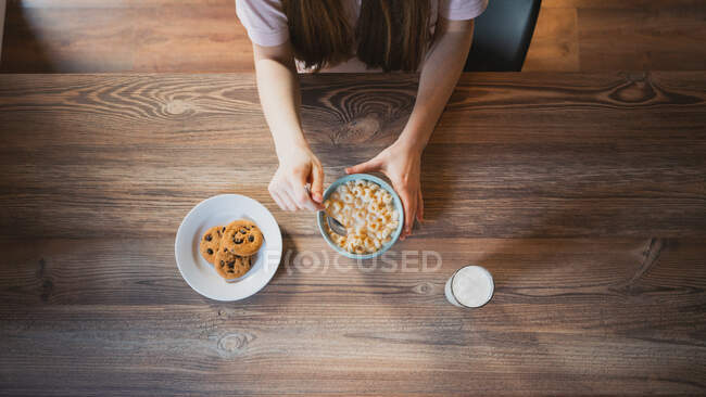 Vista superior de la cosecha femenina anónima con tazón de anillos de maíz entre deliciosas galletas de avena y vaso de leche en el interior - foto de stock