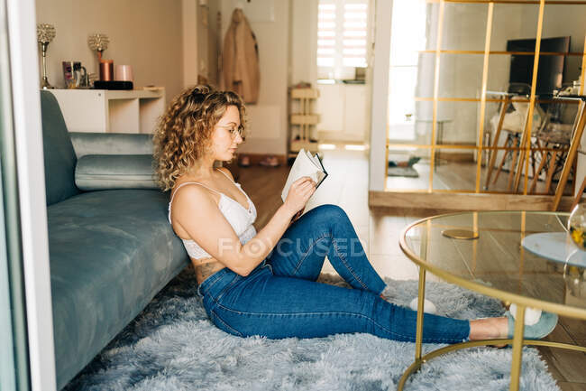 Full body side view of positive young female with curly hair in casual clothes and slippers sitting on carpet near sofa and reading interesting book during weekend at home — Foto stock