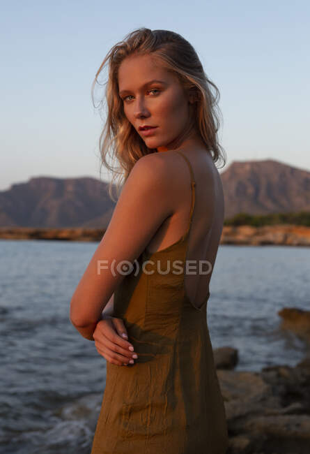 Side view of young woman standing on coast against blue waving sea at sunset and looking at camera — Stock Photo