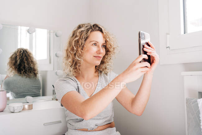 Blogger joven positiva con el pelo rubio rizado en ropa casual tomando selfie en el teléfono inteligente mientras se sienta cerca del espejo en el baño - foto de stock
