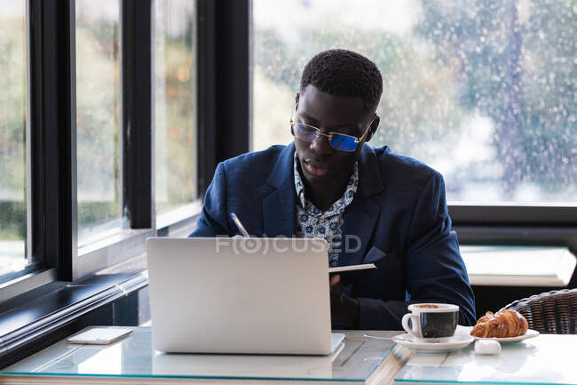 Concentrato imprenditore afroamericano di sesso maschile scrivere in blocco note mentre seduto a tavola con il computer portatile e lavorare in caffè — Foto stock