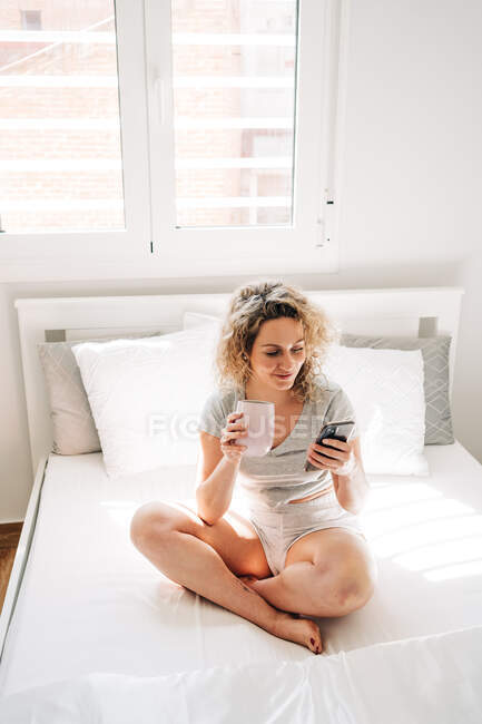 Cheerful young female with mug browsing on smartphone while sitting on comfy bed — Fotografia de Stock