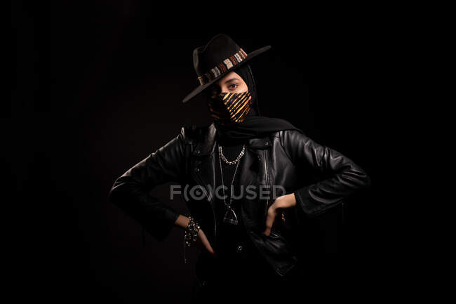 Fashionable Islamic female wearing leather jacket hijab and trendy hat standing looking at camera in dark studio during coronavirus outbreak — Stock Photo