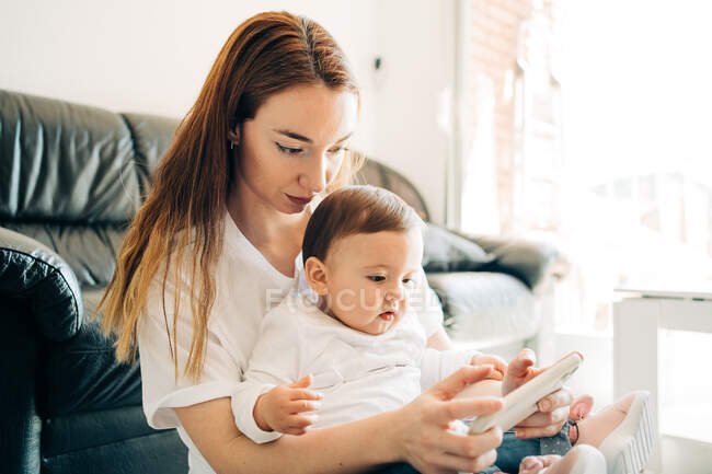Positive young mother and adorable curious baby browsing modern mobile phone together while sitting on floor in sunny living room — Stock Photo