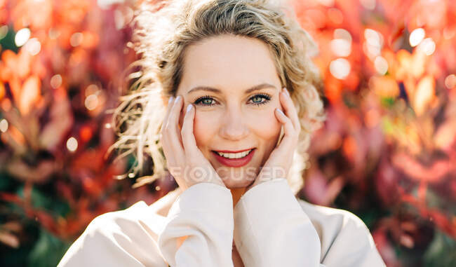 Mujer rubia feliz en vestido elegante y abrigo de pie entre los árboles y mirando a la cámara - foto de stock