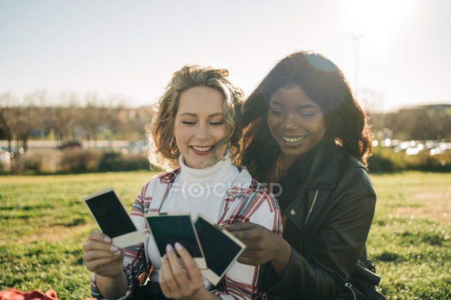 Diverse female best friends relaxing in park and looking through instant photo cards — Stock Photo