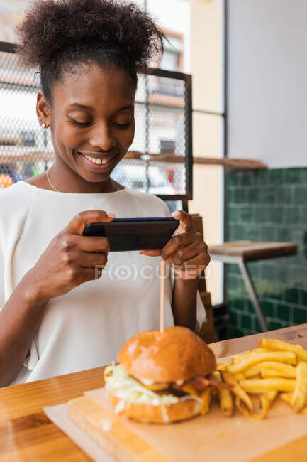Vista lateral tranquila mujer afroamericana en ropa elegante tomando fotos de deliciosas hamburguesas y papas fritas servidas en la mesa alta en el restaurante de comida rápida - foto de stock