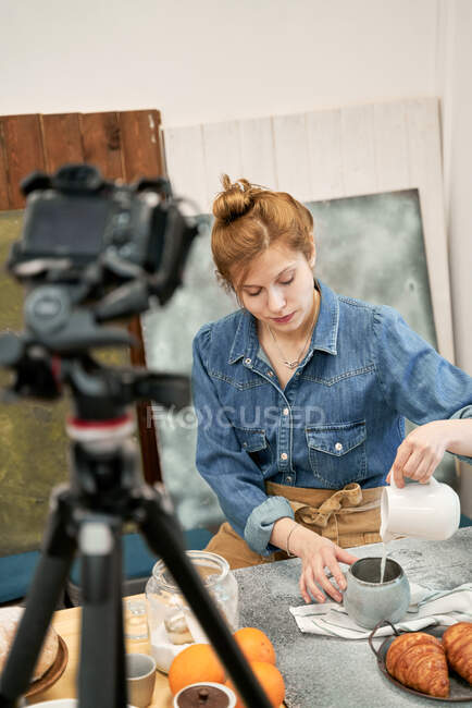 Jovem blogueira feminina derramando leite de jarro em xícara contra câmera de fotos à mesa com croissants em casa — Fotografia de Stock
