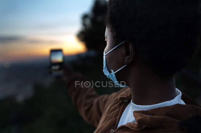 Calm young African American female in warm jacket and protective face taking photo while standing on hilltop against blurred city in twilight — Stock Photo