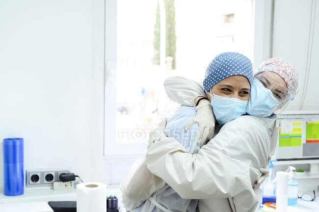 Усмішка жінок-хірургів у стерильних масках і уніформах, що обіймаються проти вікна на роботі в клініці. — стокове фото