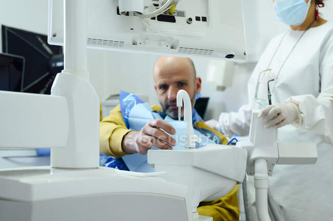 Coltiva dentista femminile irriconoscibile in maschera uniforme e sterile contro l'uomo versando acqua dal rubinetto al vetro dopo l'operazione orale — Foto stock