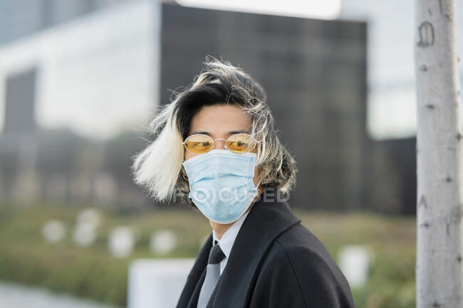 Young ethnic male executive with hair dyed in disposable mask and sunglasses looking away in city on blurred background during coronavirus outbreak — Stock Photo