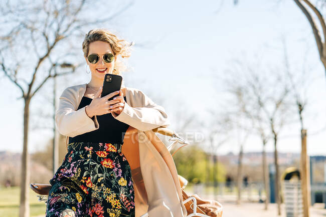 Von unten gut gelaunte Frau mit trendiger Sonnenbrille, die an sonnigen Tagen auf dem Gehweg im Garten Selbstaufnahmen mit dem Smartphone macht — Stockfoto