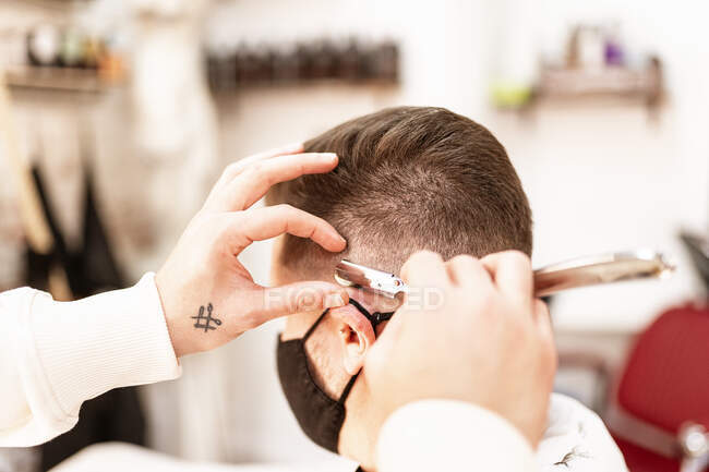 Crop анонімний перукар гоління людини в масці з прямою бритвою в перукарні на розмитому фоні — стокове фото
