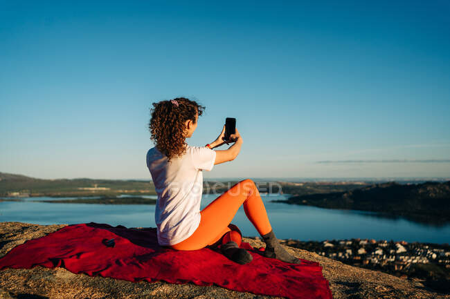 Обратный вид на неузнаваемую женщину-туристку с вьющимися волосами в повседневной одежде, делающую селфи, сидящее на одеяле на скалистом утесе над морем в солнечный день — стоковое фото