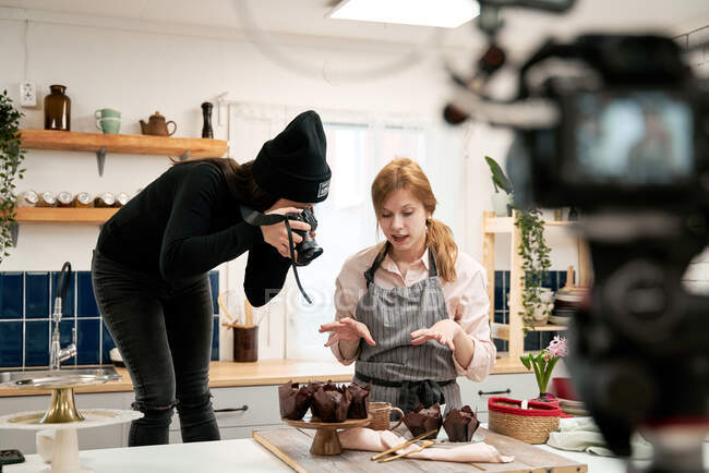 Femme méconnaissable prenant des photos de muffins au chocolat sur appareil photo numérique contre blogueur parler pendant le processus de cuisson dans la cuisine — Photo de stock