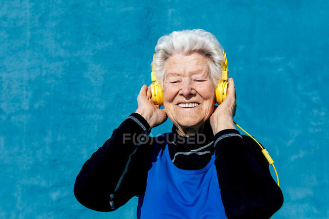 Fêmea idosa encantada com cabelos grisalhos e fones de ouvido amarelos desfrutando de músicas enquanto ouve música em fundo azul no estúdio — Fotografia de Stock