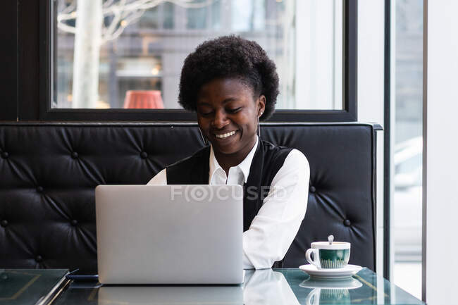 Allegro freelance afro-americana con acconciatura afro seduta a tavola con netbook mentre lavora in remoto nel caffè e distoglie lo sguardo — Foto stock