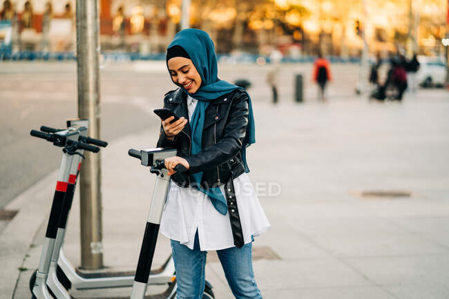 Conteúdo Feminino muçulmano em lenço de cabeça tradicional usando o aplicativo no smartphone e exibição de digitalização para desbloquear scooter elétrico moderno estacionado na rua da cidade — Fotografia de Stock