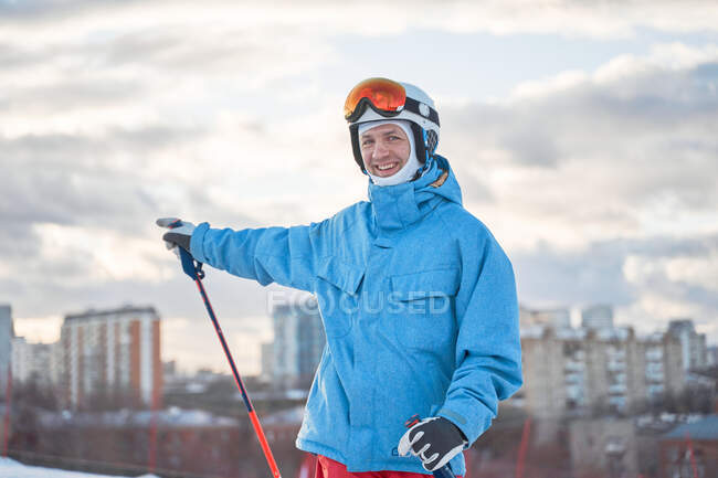 Esquiador macho feliz em sportswear quente em pé na encosta da colina nevada no parque da cidade de inverno — Fotografia de Stock