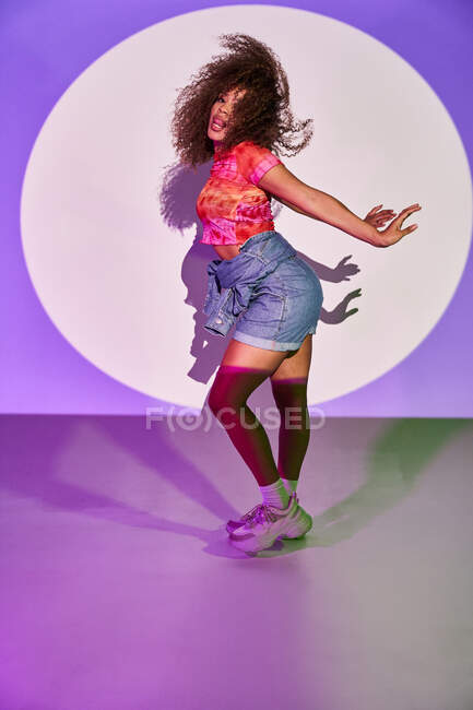 Vista lateral da dançarina afro-americana em shorts dançando olhando para a câmera com a língua para fora enquanto estava em luzes de néon no estúdio — Fotografia de Stock