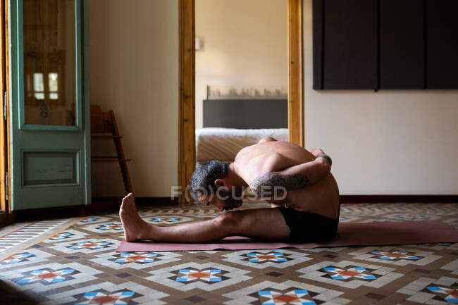 Vista lateral do macho com tronco nu praticando ioga no tapete em Marichyasana em casa — Fotografia de Stock