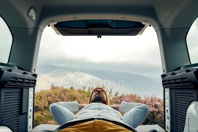 Jovem campista masculino em outerwear deitado na van com os olhos fechados desfrutando de vista panorâmica das terras altas — Fotografia de Stock