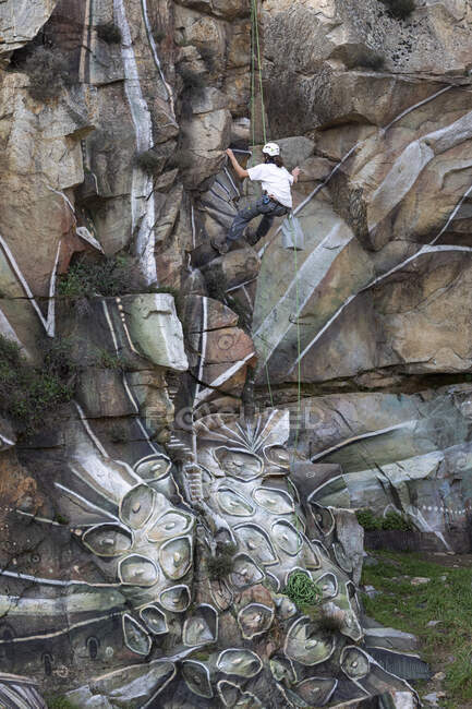 Visão traseira corpo inteiro do pintor com tinta spray fazendo graffiti pendurado na corda em encosta rochosa íngreme — Fotografia de Stock
