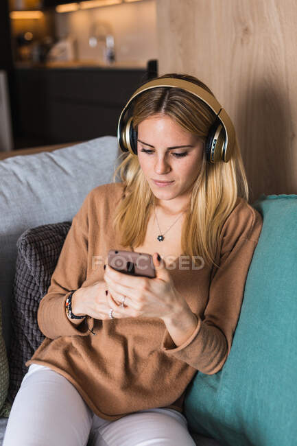 Mujer joven sentada en el sofá y disfrutando de la música en los auriculares mientras mira la pantalla del teléfono inteligente - foto de stock