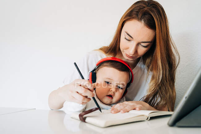 Positive junge Mutter mit Bleistift lehrt entzückend fleißiges Baby mit Kopfhörern, Tagebuch zu schreiben, während es zusammen am Schreibtisch im hellen Raum sitzt — Stockfoto