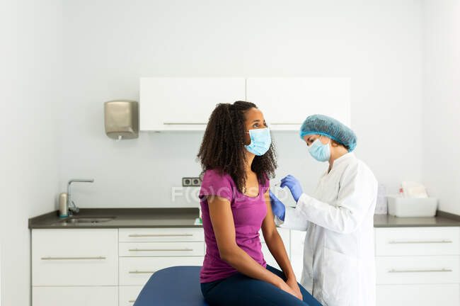 Специалистка-женщина в защитной форме, латексных перчатках и маске для лица, прививающая афроамериканку в клинике во время вспышки коронавируса — стоковое фото