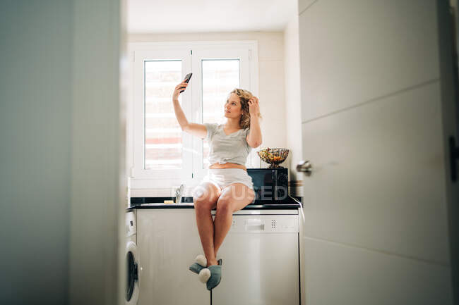 Cuerpo completo joven contenido femenino en ropa doméstica casual tomando selfie en el teléfono inteligente mientras está sentado en el mostrador de la cocina y tocando el pelo rizado - foto de stock
