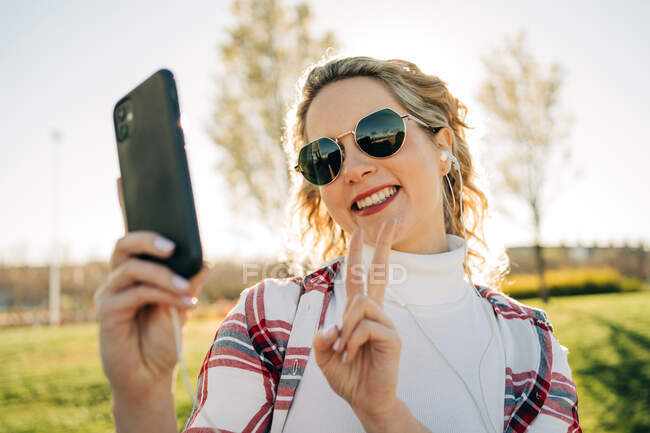 Mujer positiva en auriculares tomando autorretrato del teléfono móvil y mostrando el gesto de dos dedos mientras está de pie en el parque al atardecer - foto de stock
