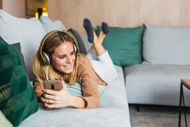 Радостная женщина лежит на диване и наслаждается музыкой в наушниках, отворачиваясь, используя смартфон — стоковое фото
