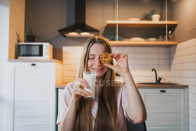 Contenido joven hembra con los ojos cerrados y vaso de leche con galleta de avena para el desayuno en casa - foto de stock