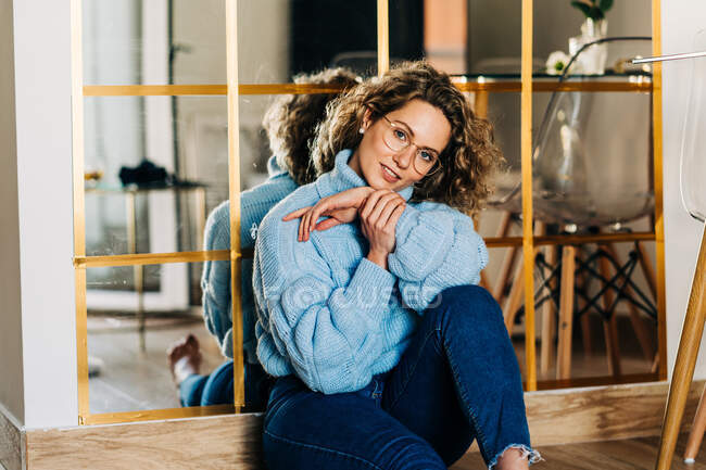Stylish young female with curly blond hair in trendy warm sweater and eyeglasses sitting on floor near mirror and looking at camera — Foto stock