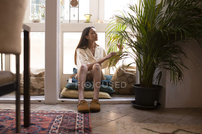 Jovem fêmea calma em roupas casuais removendo folhas secas de plantas de casa envasadas exuberantes enquanto sentado em travesseiros perto da janela em casa — Fotografia de Stock