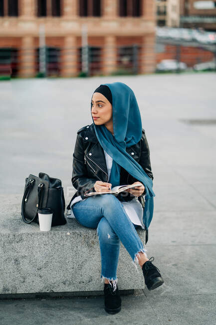 Thoughtful Muslim female in hijab writing in diary while sitting on city street and looking away — Stock Photo