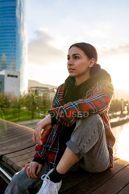 Pensive ethnic female sitting with crossed legs in town while looking away in sunlight — Stock Photo